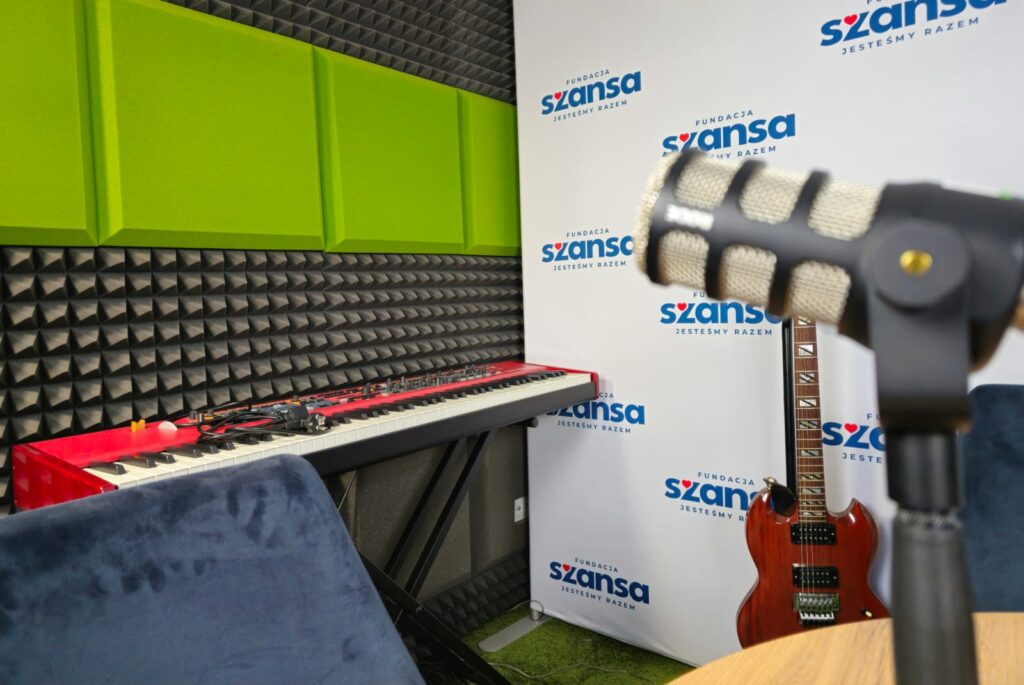 Czerwone pianino cyfrowe pod ścianą z zielonych i czarnych paneli akustycznych; obok gitara elektryczna i mikrofon na statywie