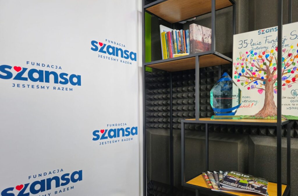 Kącik ekspozycyjny: regał z książkami, szklana statuetka i plakat 35-lecia Fundacji Szansa z drzewem z kolorowych odcisków palców